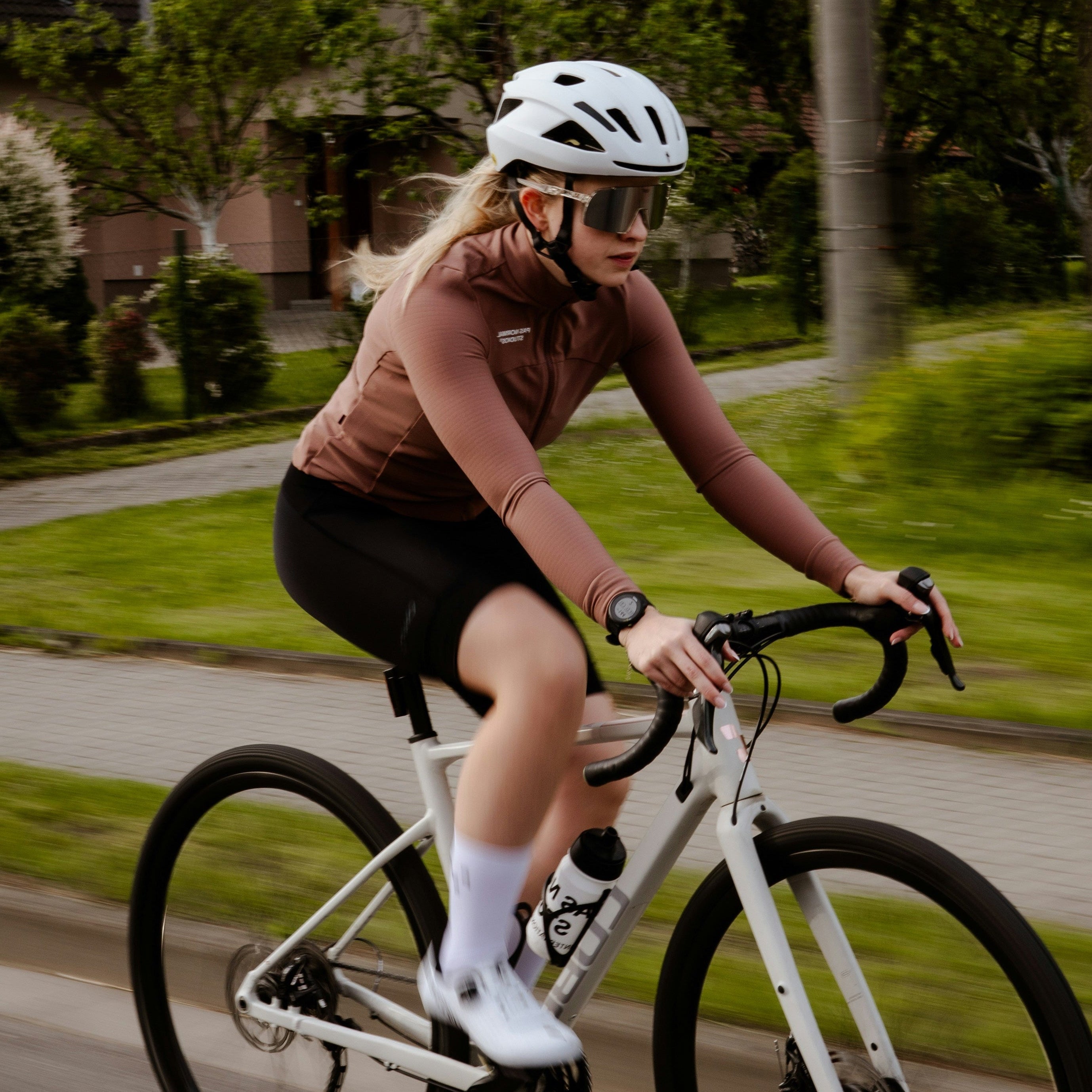 A woman riding a white road/gravel bicycle on a suburban road, wearing a white helmet, brown long-sleeve jersey, black shorts, and white socks.