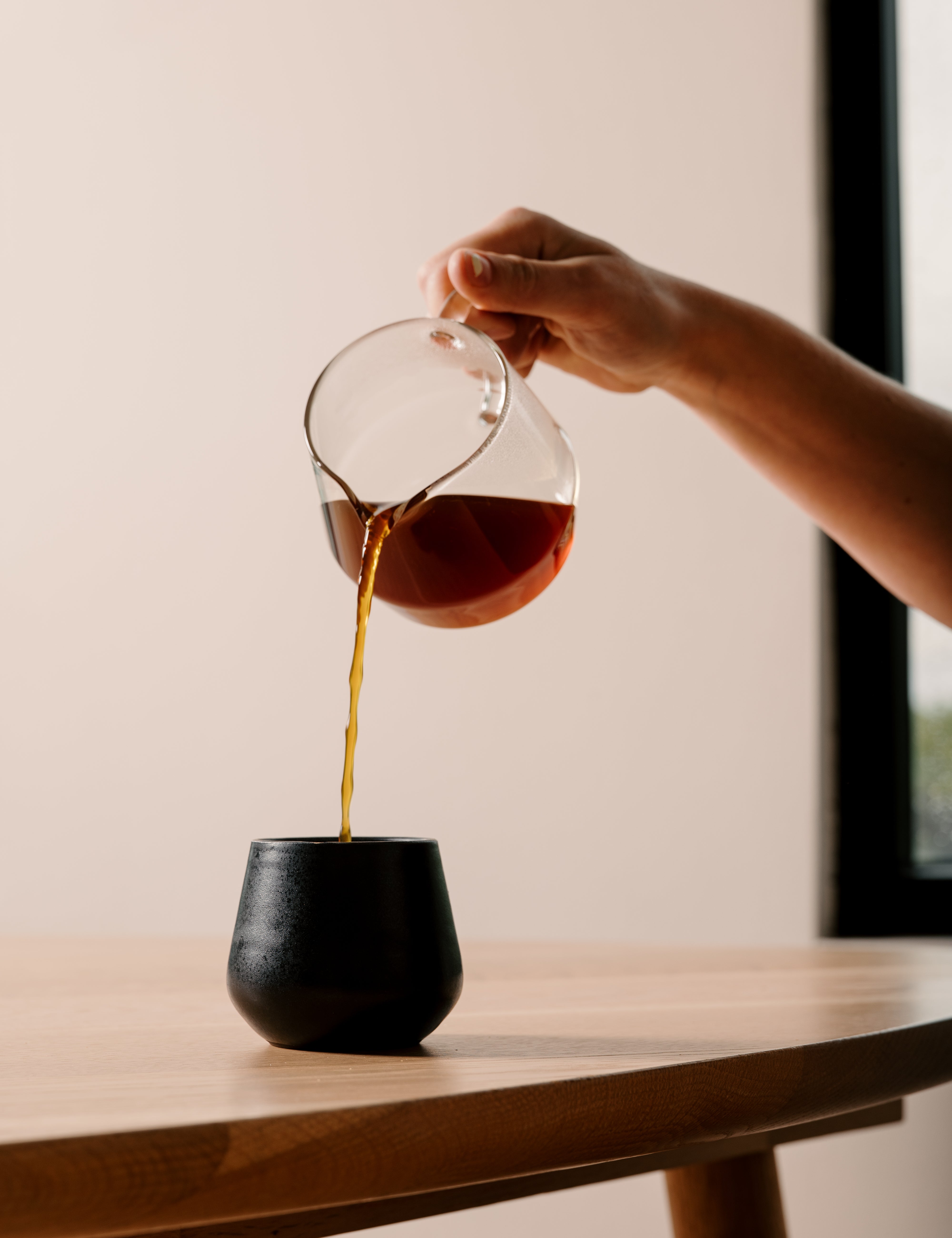 A hand pours dark coffee from a clear glass pour-over carafe into a matte black cup on a light wood table.